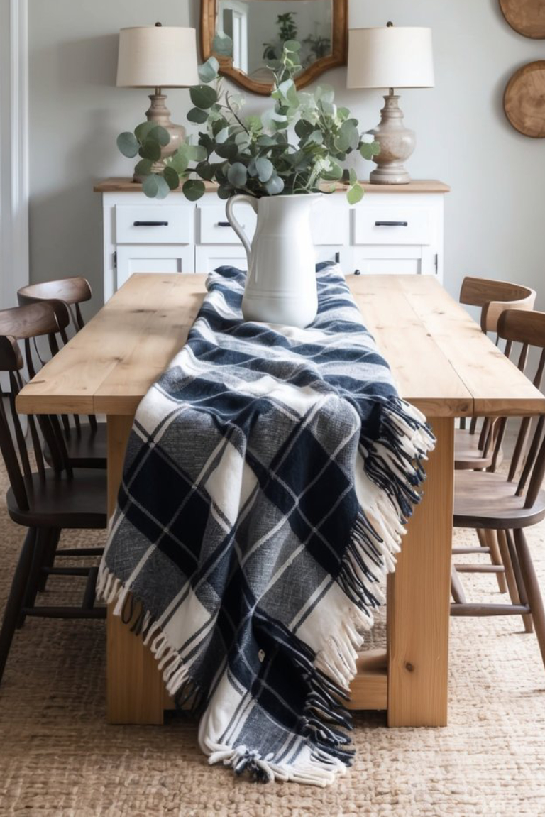 A dining table with a light wood surface, covered by a navy and white plaid blanket, topped with a white pitcher filled with eucalyptus leaves. The scene includes wooden chairs and a light-colored rug underneath, along with decorative wall plates and lamps in the background.