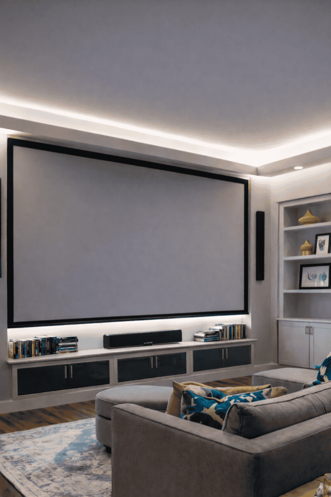 A modern living room featuring a large wall-mounted projection screen with a black frame, a low-profile media center below it with a soundbar and a collection of books, and an inviting gray sectional sofa adorned with colorful throw pillows, all set against a softly lit backdrop with recessed lighting.